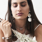 A woman with hey eyes closed holding plants. She is wearing pendants and a chain necklace with beads.