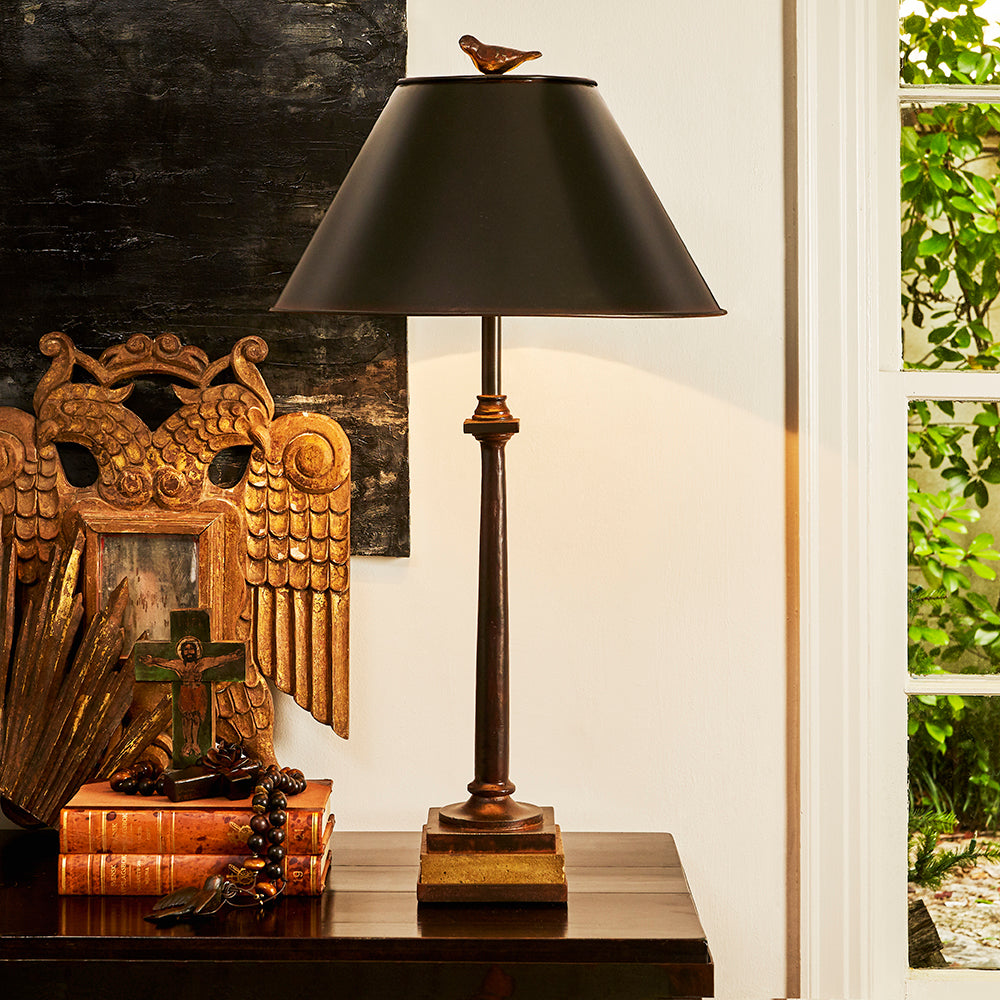 A wax cast bird in an iron lamp. On top of a table next to a window and some books with a cross.