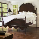A white bed with a iron frame. To its side is a lamp with a dark shade and two windows.