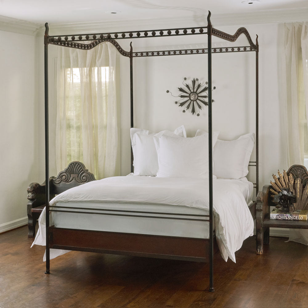 A canopy bed with white pillows and sheets- Inside a room with white walls and wood floor.