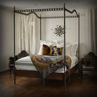 A canopy bed with white pillows and sheets- Inside a room with white walls and wood floor. On top of the bed is a mantel with faces and a pillow with the face of the virgin.