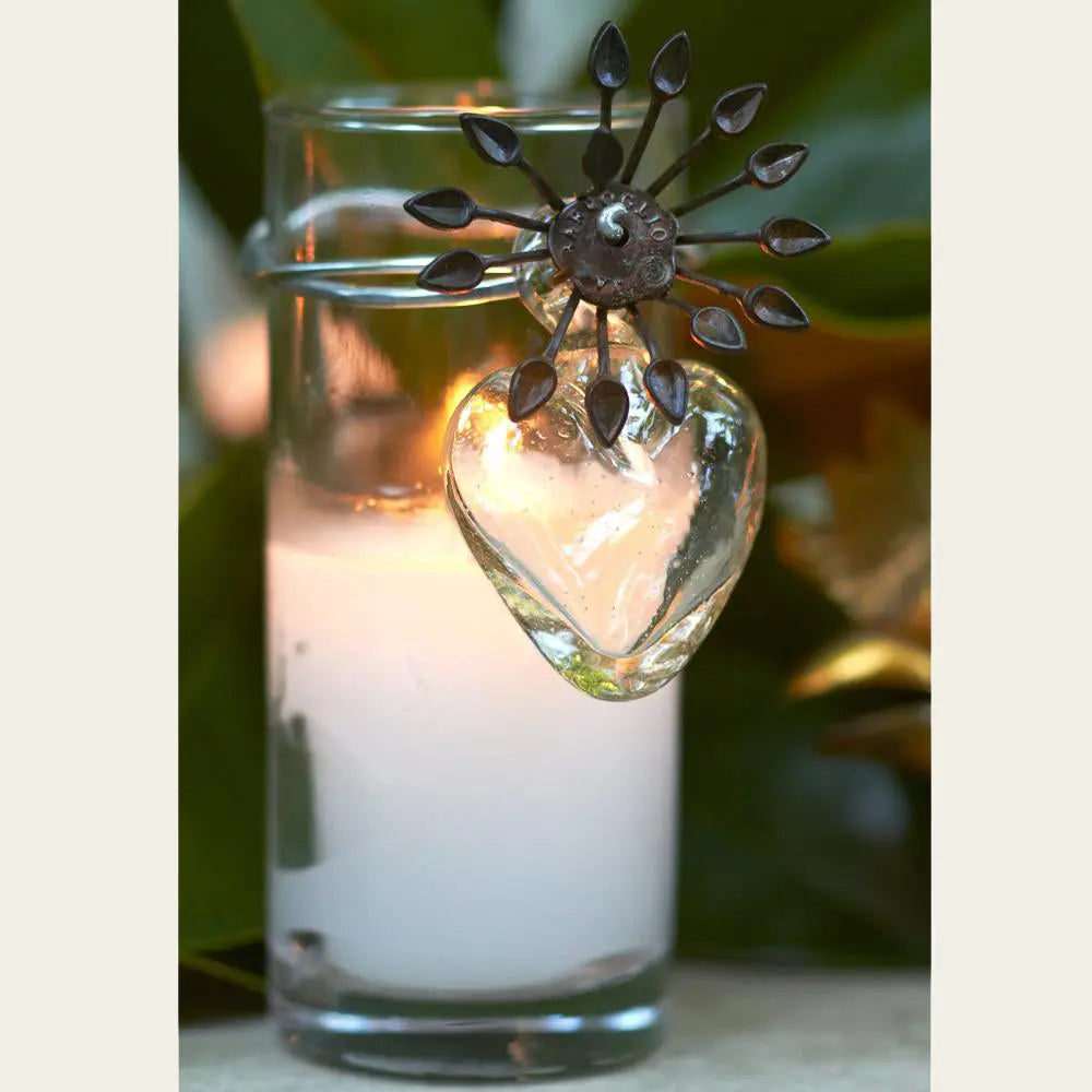 A poured candle in glass votive with mouth blown glass heart, iron flower and rafta. Outside background.