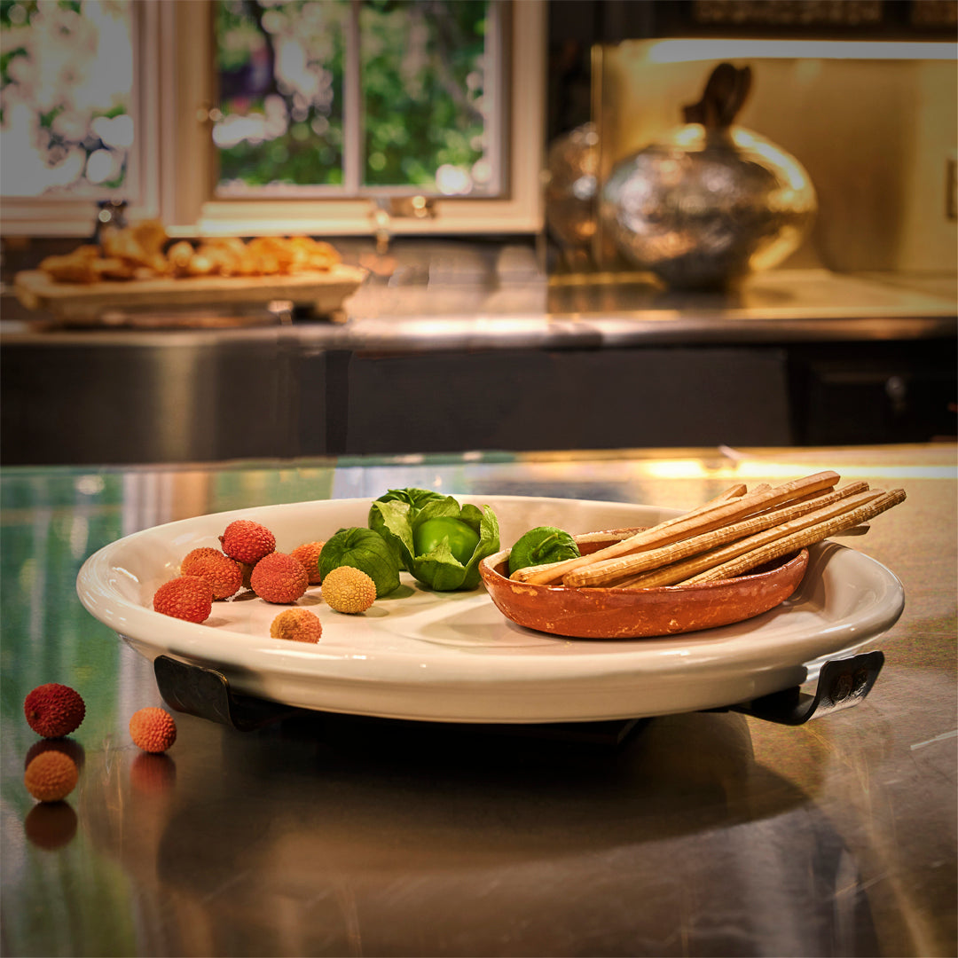 A iron base holding a white ceramic plate with some fruits, green vegetables and a brown plate with breadsticks. 