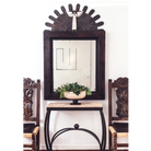A forged iron table with a carved stone top. with a pot with plants on top. It is positioned between two chairs and underneath a mirror.