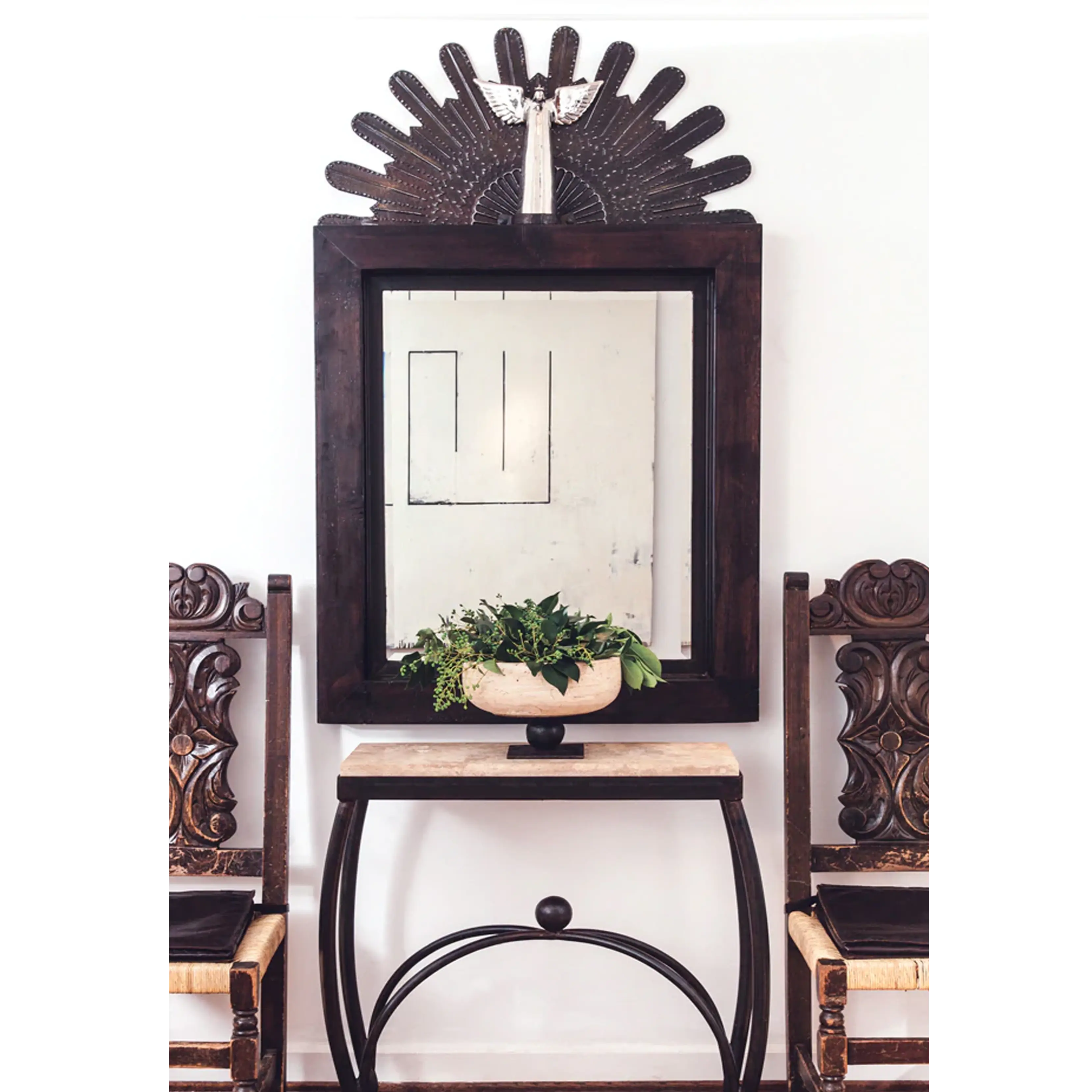 A forged iron table with a carved stone top. with a pot with plants on top. It is positioned between two chairs and underneath a mirror.