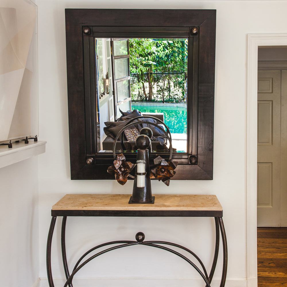 A forged iron console table with carved  stone top. On top of the table is an iron angel holding a candle and a mirror reflecting a sofa and a pool. Everything is against a white wall.