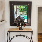 A forged iron console table with carved  stone top. On top of the table is an iron angel holding a candle and a mirror reflecting a sofa and a pool. Everything is against a white wall.