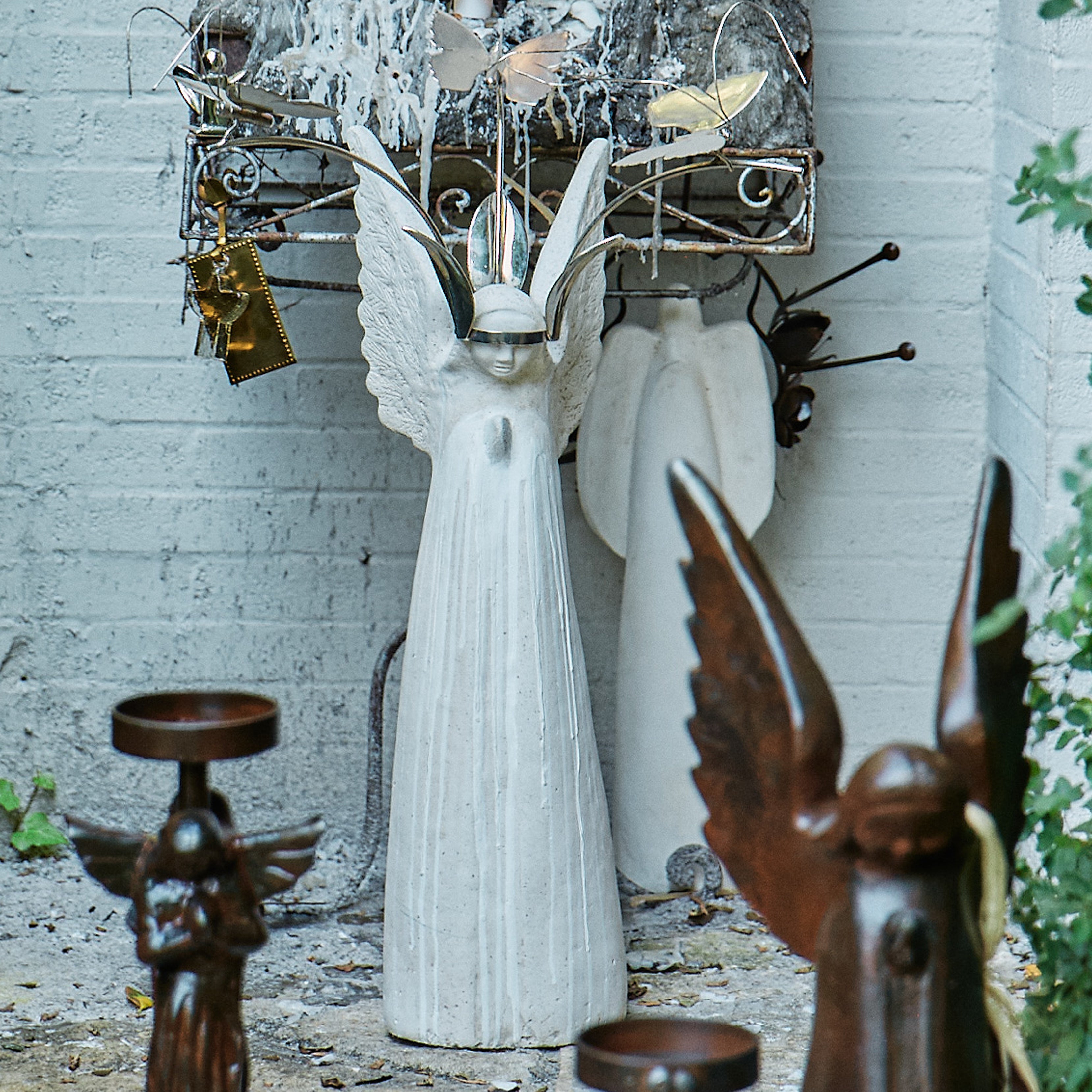A white stone angel with nickel butterfly crown and white blessing beads with flowers. It is resting against a white brick wall. There are two iron angels in front.