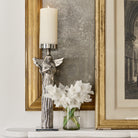 Nickel plated wax cast iron double angel on stand with a candle. Next to a glass vase with white flowers on top of a white background.