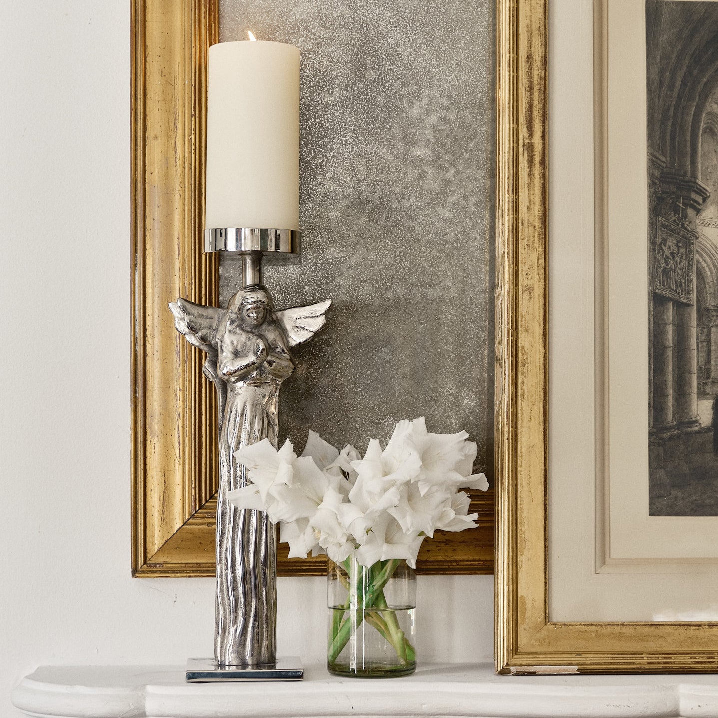Nickel plated wax cast iron double angel on stand with a candle. Next to a glass vase with white flowers on top of a white background.