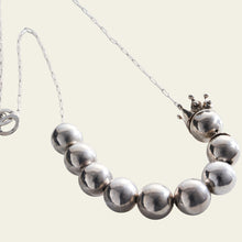 A necklace with nine silver spheres in a delicate sterling cable chain necklace. A ruler's crown is set askew. Plain white background.