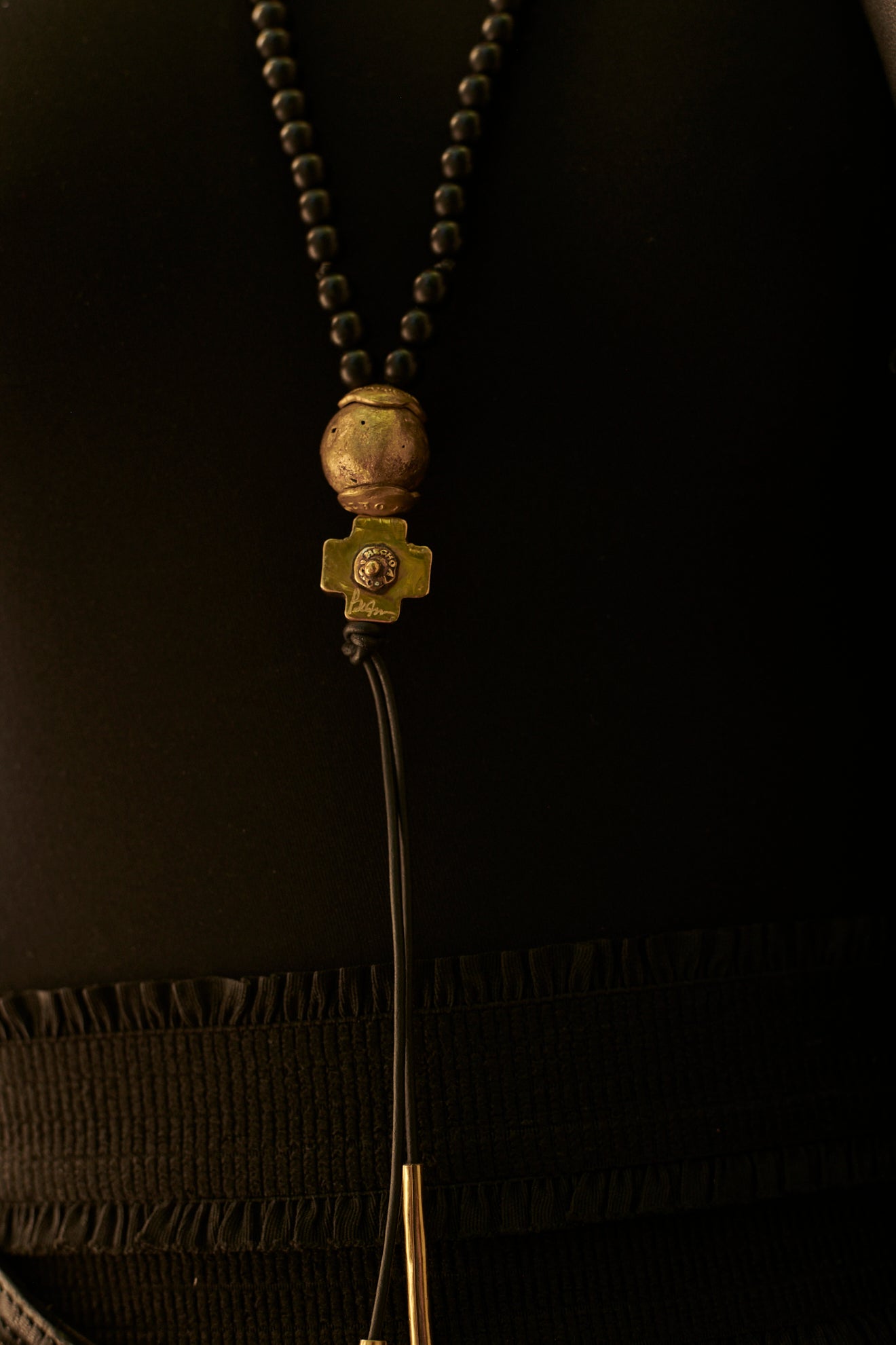 A necklace with black beads and at the end a bronze ball and a bronze cross followed by two black leather strings with two bronze cylinders or topers. Black background.