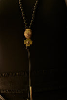 A necklace with black beads and at the end a bronze ball and a bronze cross followed by two black leather strings with two bronze cylinders or topers. Black background.