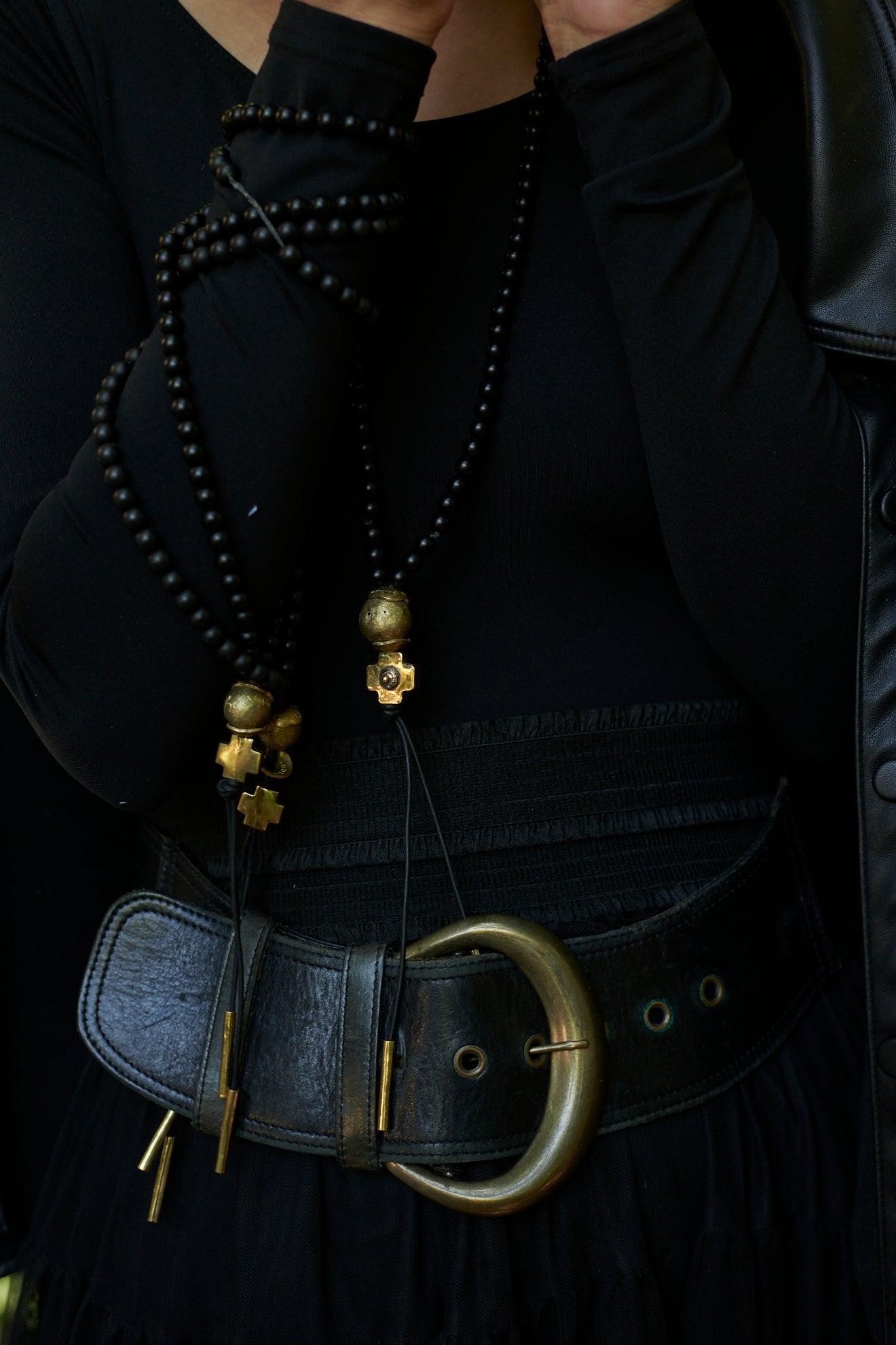 Several  necklaces with black beads and at the end a bronze ball and a bronze cross followed by two black leather strings with two bronze cylinders or topers. Neckalces wrap around a woman's wrist. 