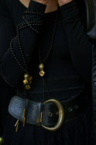 Several  necklaces with black beads and at the end a bronze ball and a bronze cross followed by two black leather strings with two bronze cylinders or topers. Neckalces wrap around a woman's wrist. 
