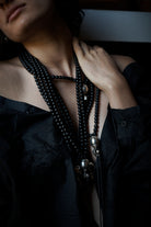 A woman resting her hand on her neck while wearing a black shirt and several  necklaces with black beads and at the end a silver ball and a silver cross followed by two black leather strings with two silver cylinders or topers. 