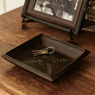 A iron valet tray with embossed cross and laurel with three keys in it. On top of a wood surface in front of a photo frame.