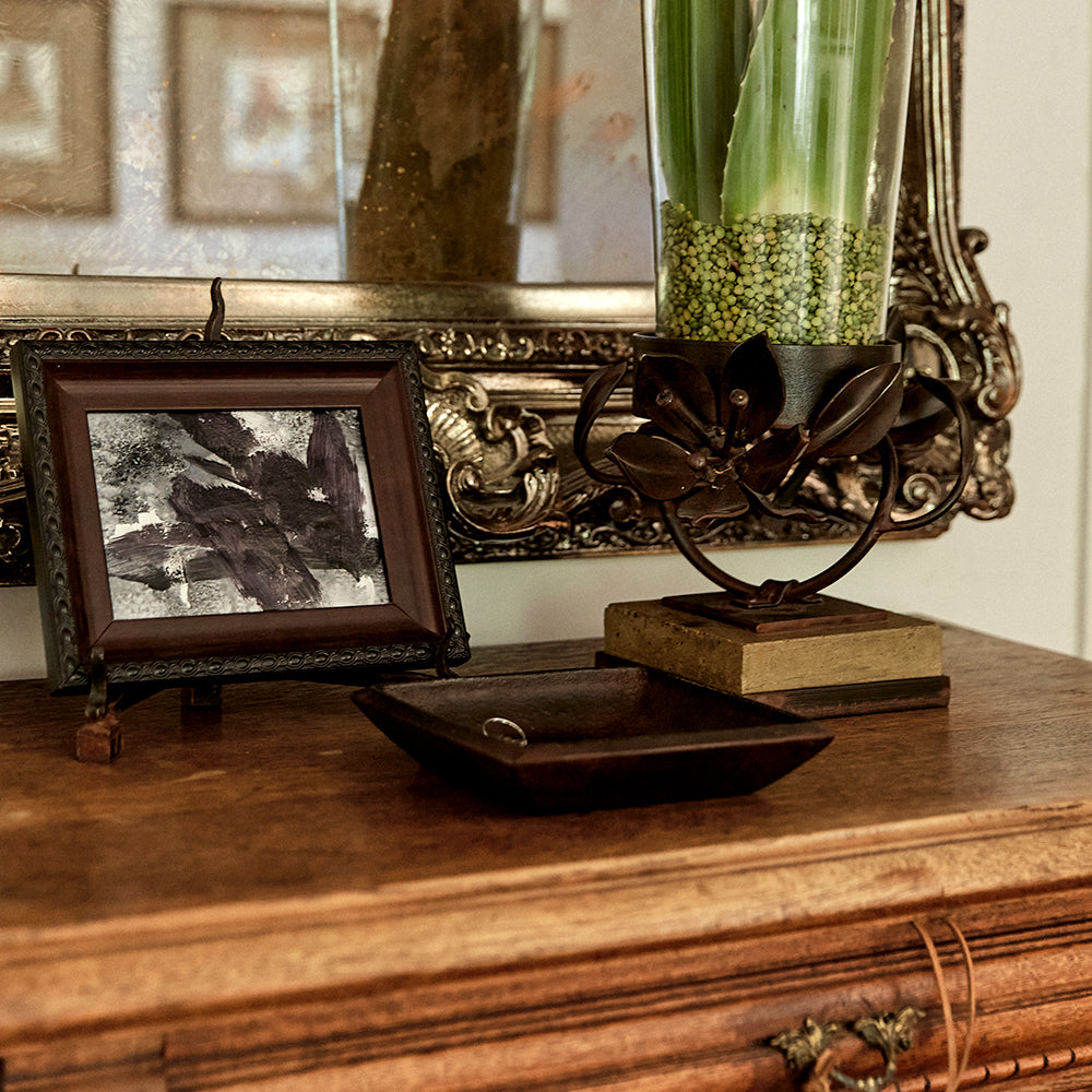 A wood furniture with a picture frame, a iron tray and a iron vase holding a glass with green leaves and seeds.