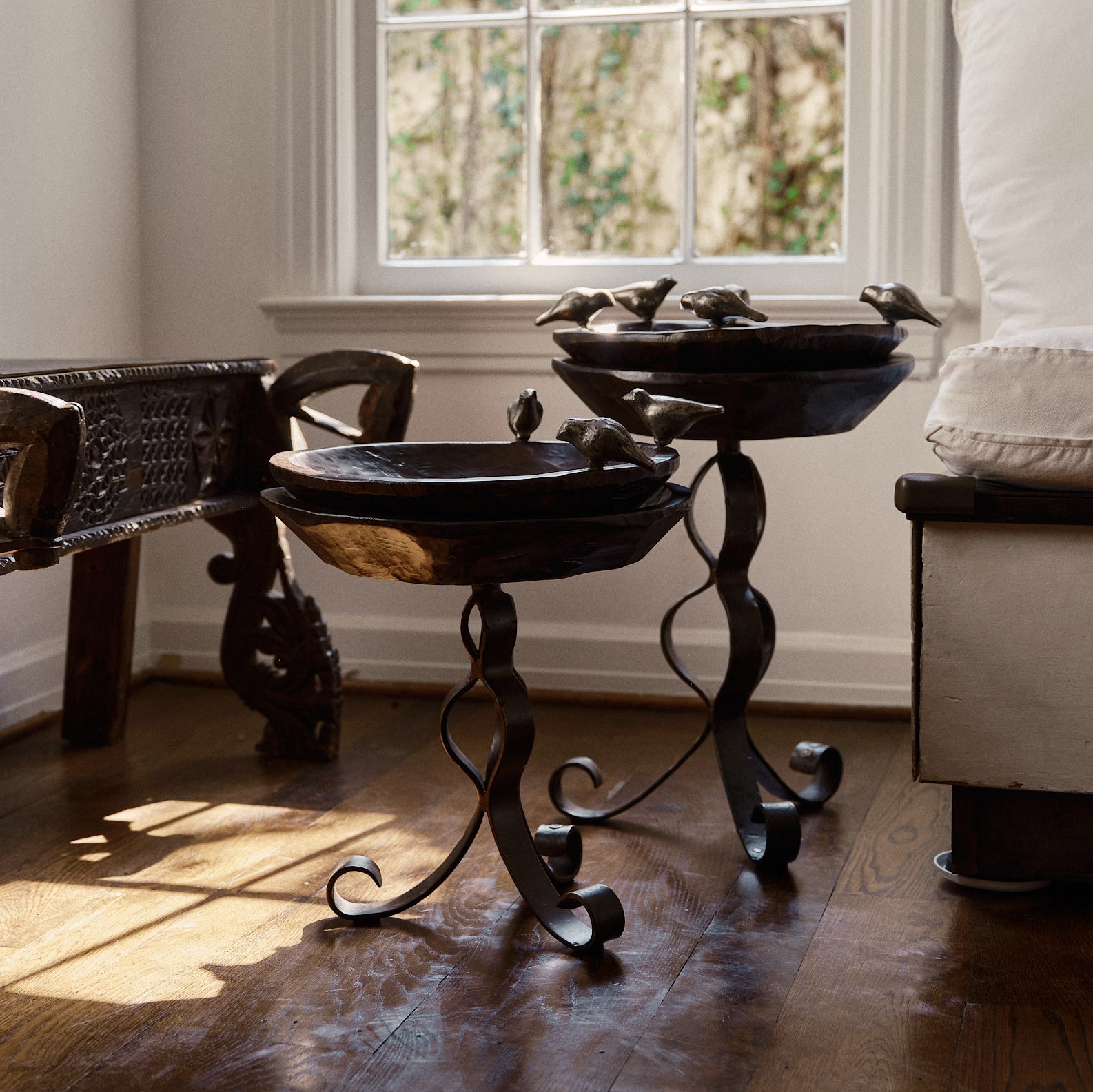 Two hand carved 16" primitive wood bowl with five wax cast iron birds on top of iron tables. In front of a window.