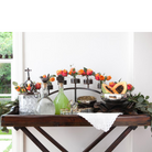 A candelabra with hand forged base and 9 glass vessels with orange flowers in each. Surrounded by  glass bottles with ice and drinks, drinking glasses and owls with seeds and fruits. Everything is on top of a table against a white wall and a window.