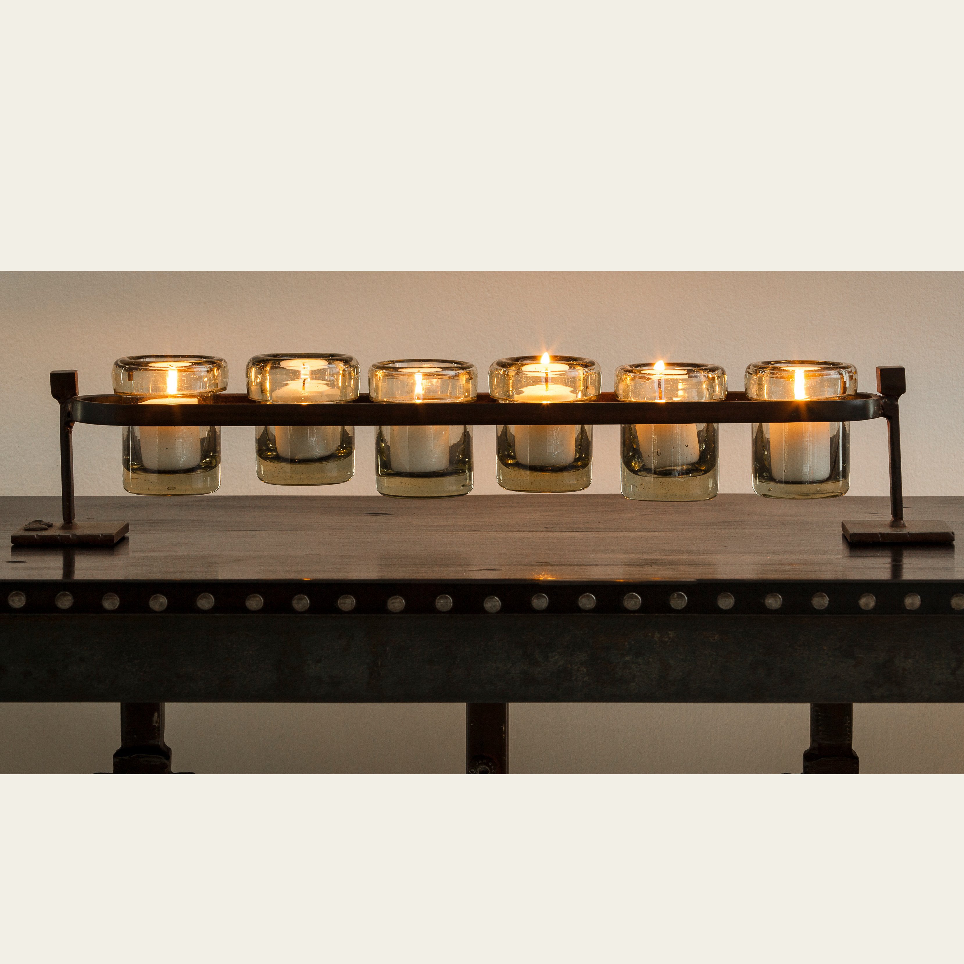 Lit candles inside a wax cast railroad with six mouth blown glass votive holders on forged stand