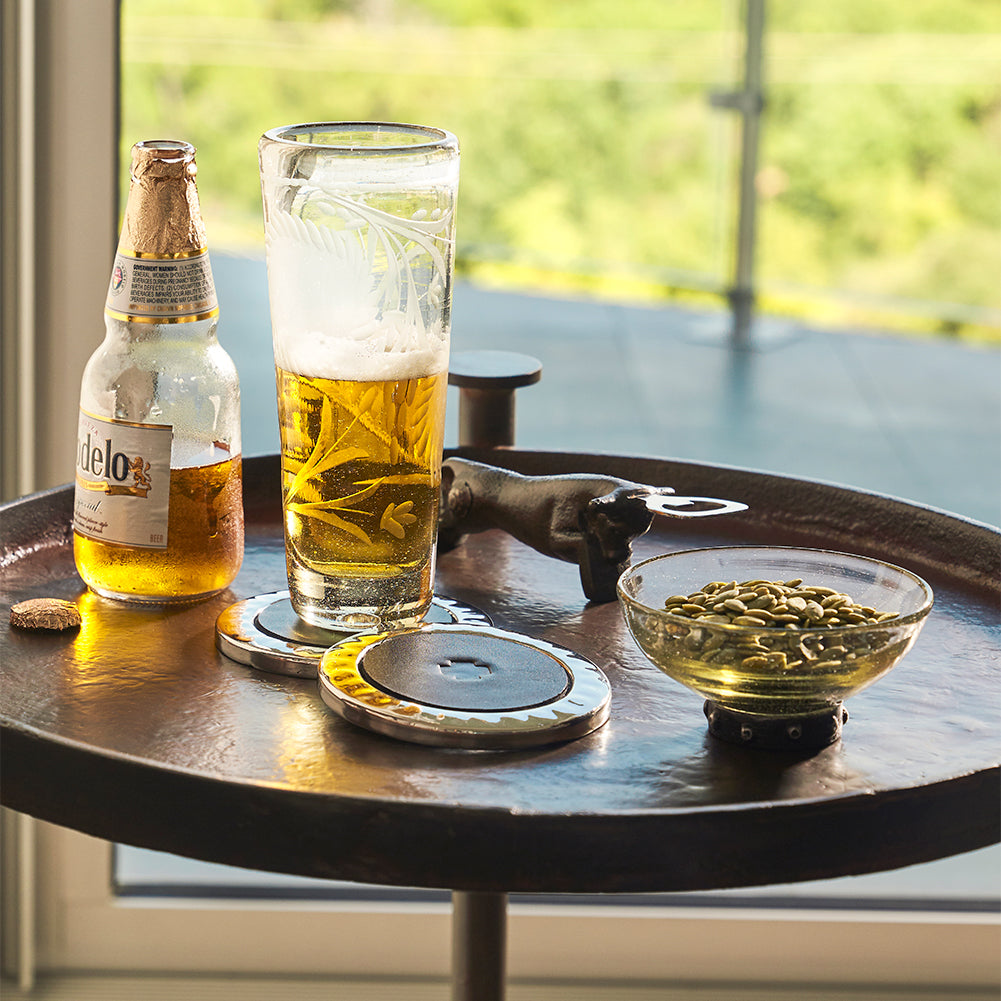 A iron round table holding a glass with beer, a beer,  two coasters, a small bowl with seeds and a bottle opener in the shape of a bull. the background is a widnow.
