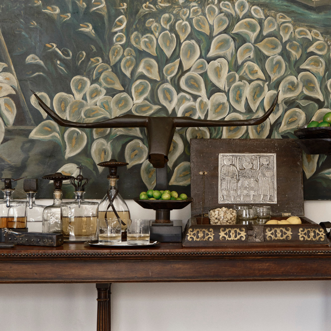 A hand-shaped iron sculpture with the shape of a bull's head with long horns. On top of a wood table with several bottles with liquid, lemons and a box with some food. On the background is a painting with white flowers.