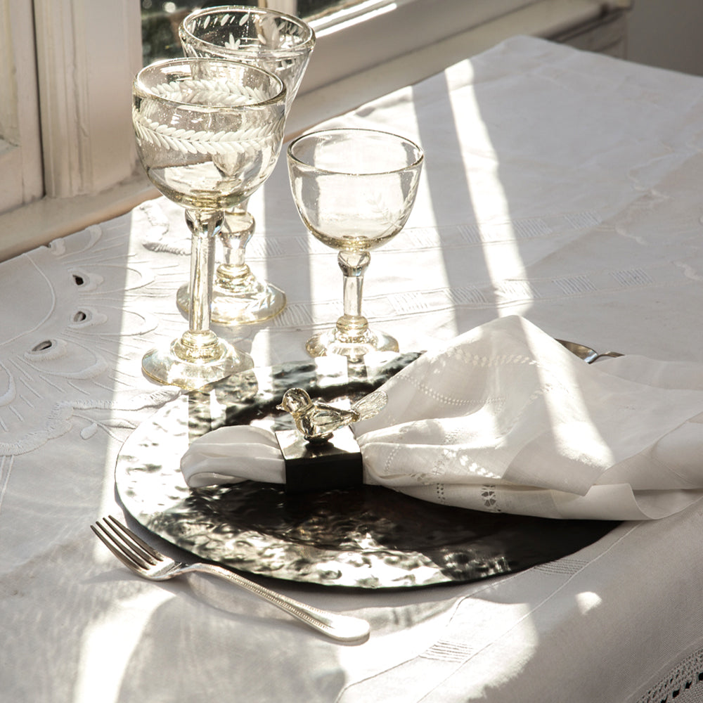 A table with a white fabric. On top is a tray with a white fabric napkin and a crystal duck, a silver fork and three clear glass cups.