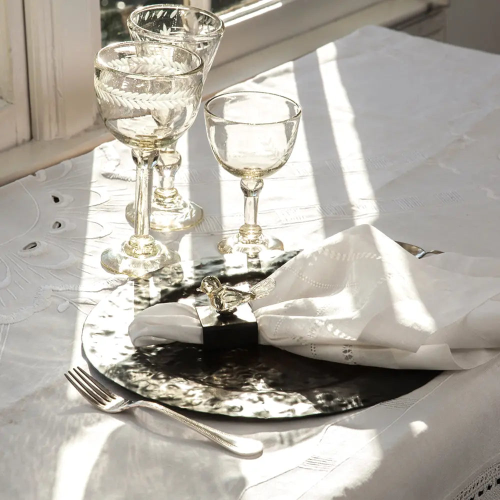 A white fabric  with three glass cups on top and a iron charger with a white napkin and a fork.