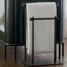 A forged iron blanket or towel rail with a white blanket. Resting against a black surface. Wooden floor.