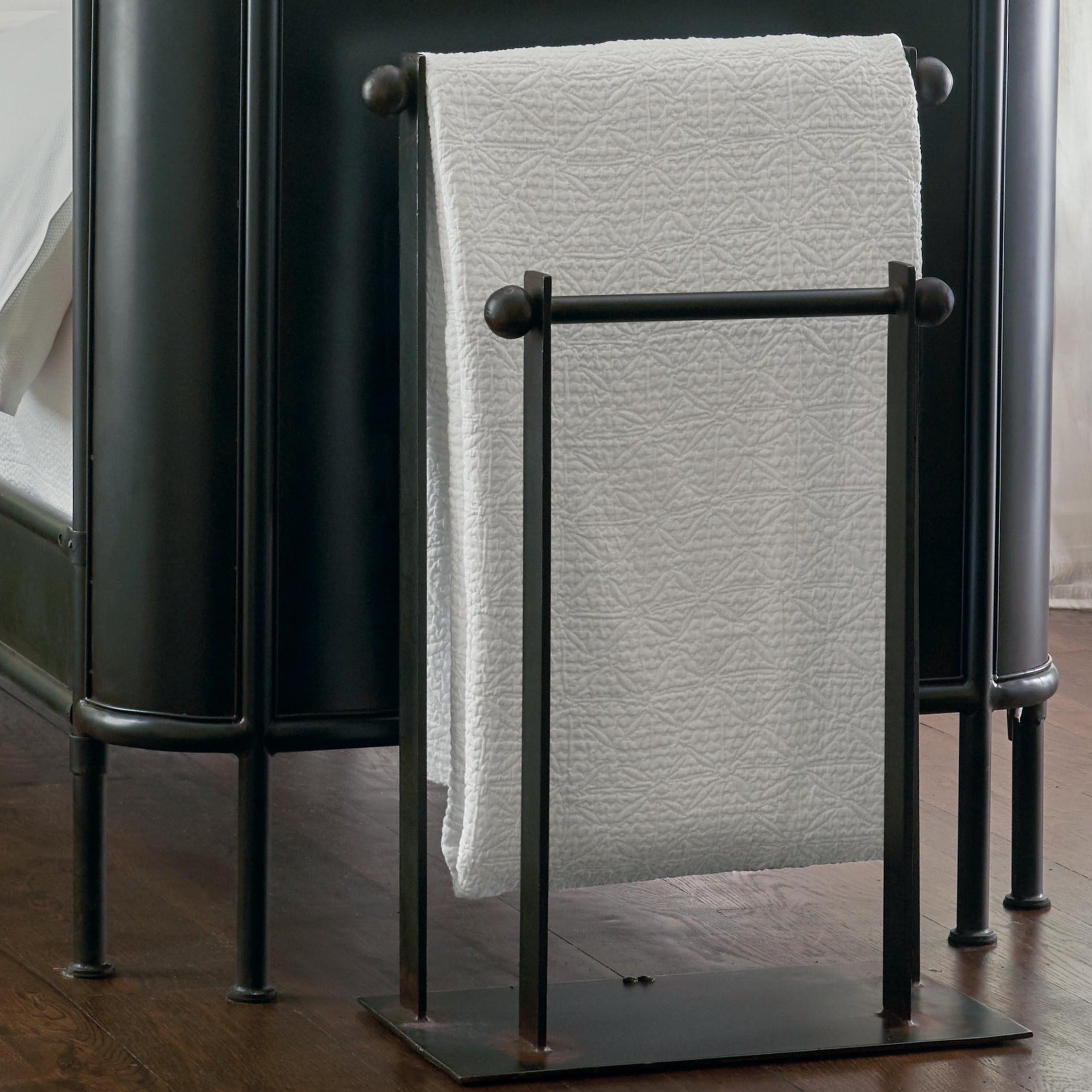 A forged iron blanket or towel rail with a white blanket. Resting against a black surface. Wooden floor.