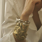 A woman wrist with a bronze cuff with a rose engraved. Three silver and bronze bracelets with silver studs on each end. The background is the white button up shirt the woman is wearing.