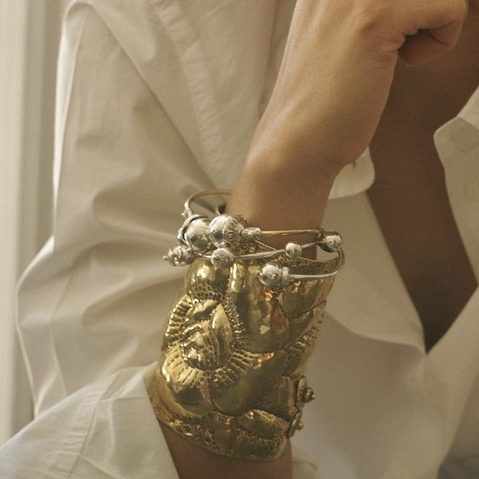 A woman wrist with a bronze cuff with a rose engraved. Three silver and bronze bracelets with silver studs on each end. The background is the white button up shirt the woman is wearing.