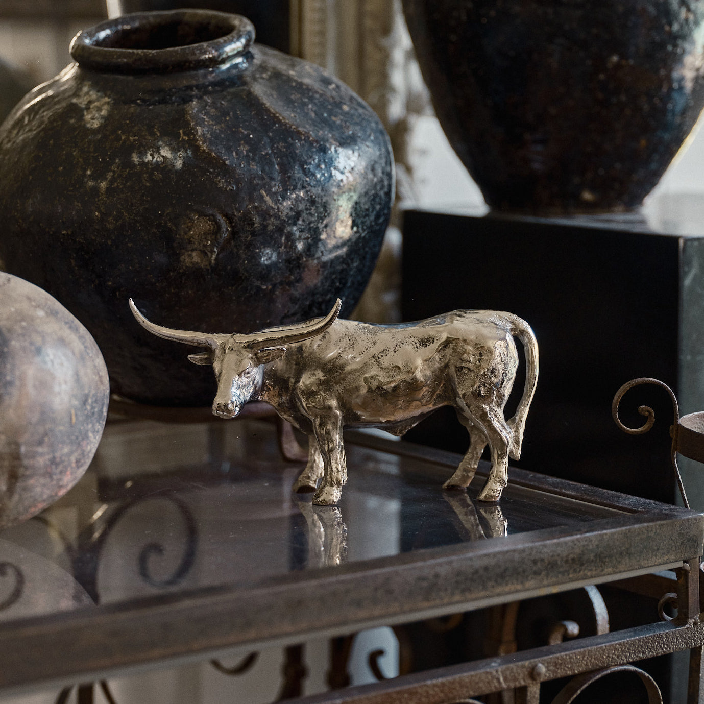 A  nickel plated longhorn bull  iron sculpture on top a glass table. Behind is a black vase.