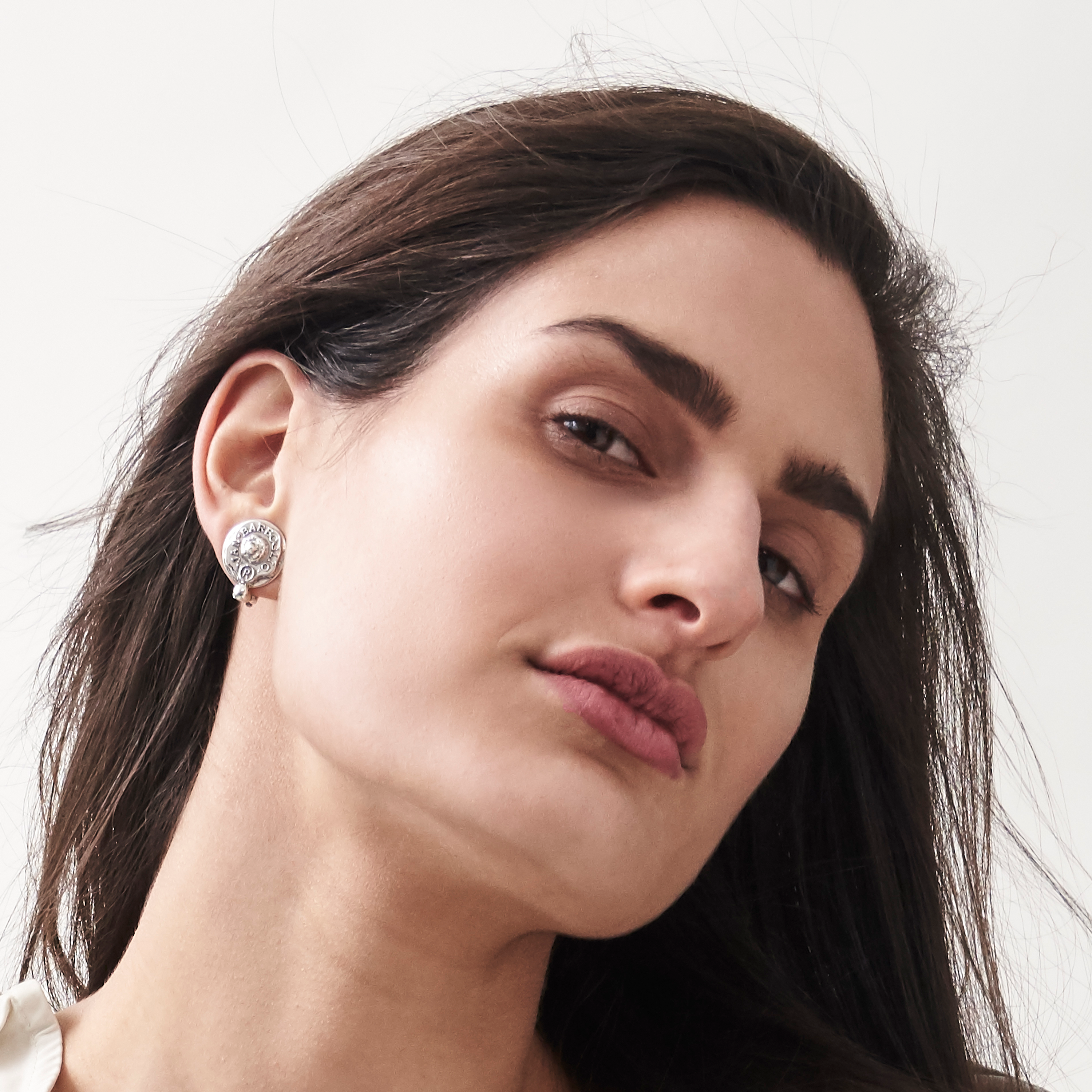 A brunette woman tilting her head wearing a silver earring with a bead. Plain background.