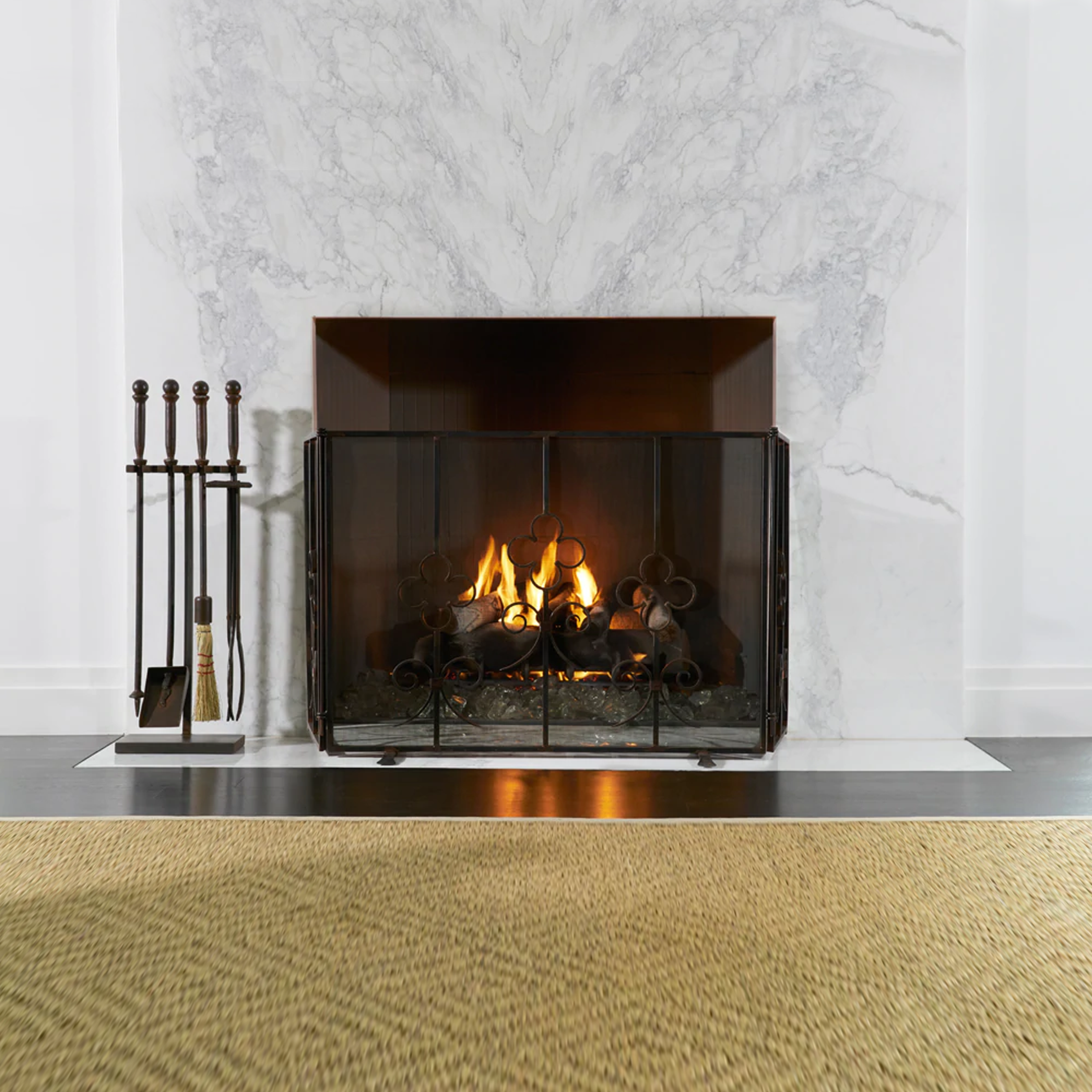 A lit white chimney with a fire screen and to the left a set of tools. A light brown rug is in front.