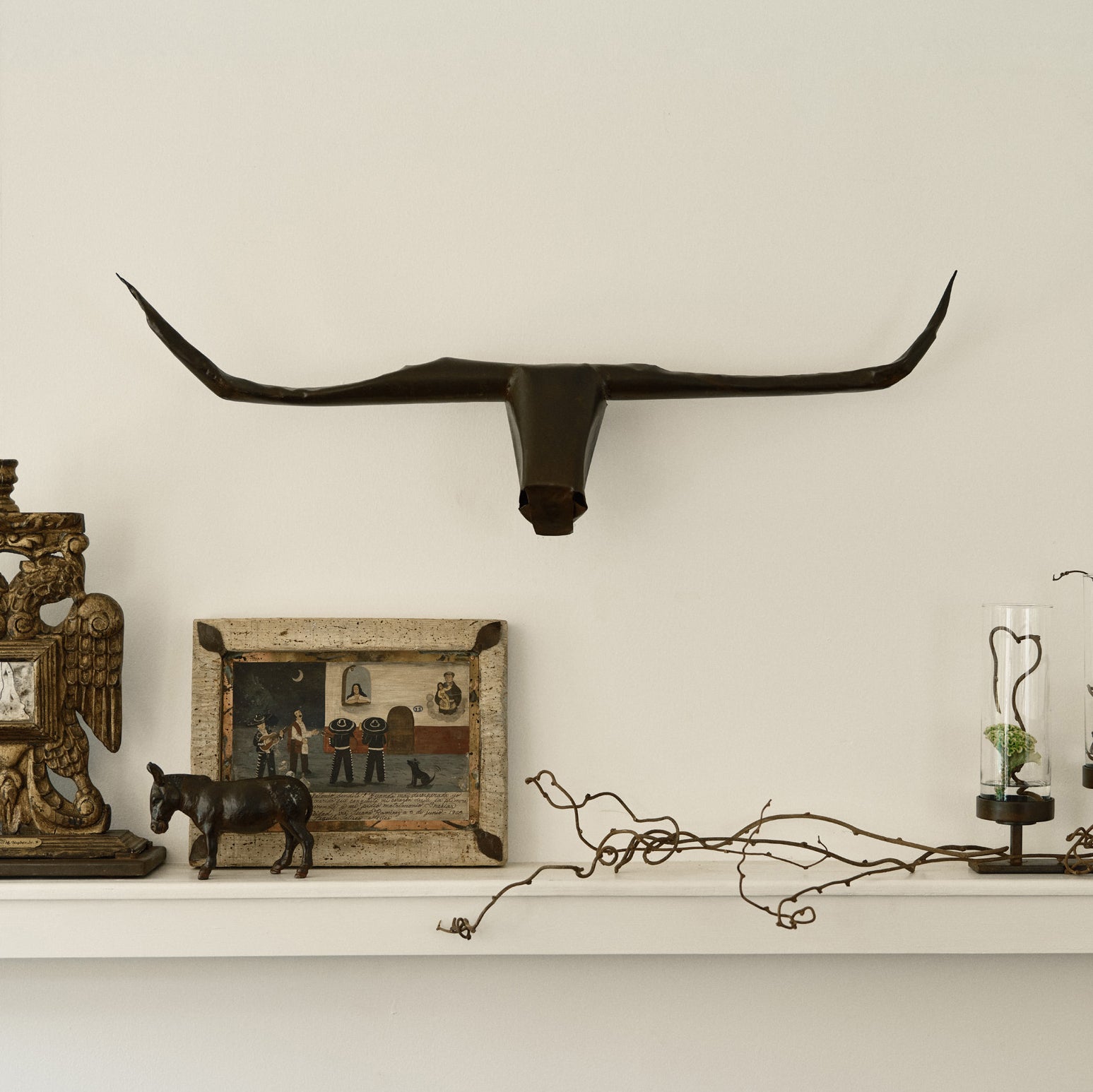 A hand-shaped iron sculpture with the shape of a bull's head with long horns hanging on a white wall. Underneath it is a branch, an iron donkey, A picture and other objects.