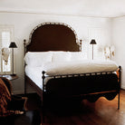 A white bed with a iron frame. On both sides is a dark lamp and cow skulls. Wood floor and white wall.