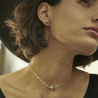 Part of a  short haired woman's face  looking up while wearing a black hat.She is wearing a delicate silver necklace with a cross and a silver earring.