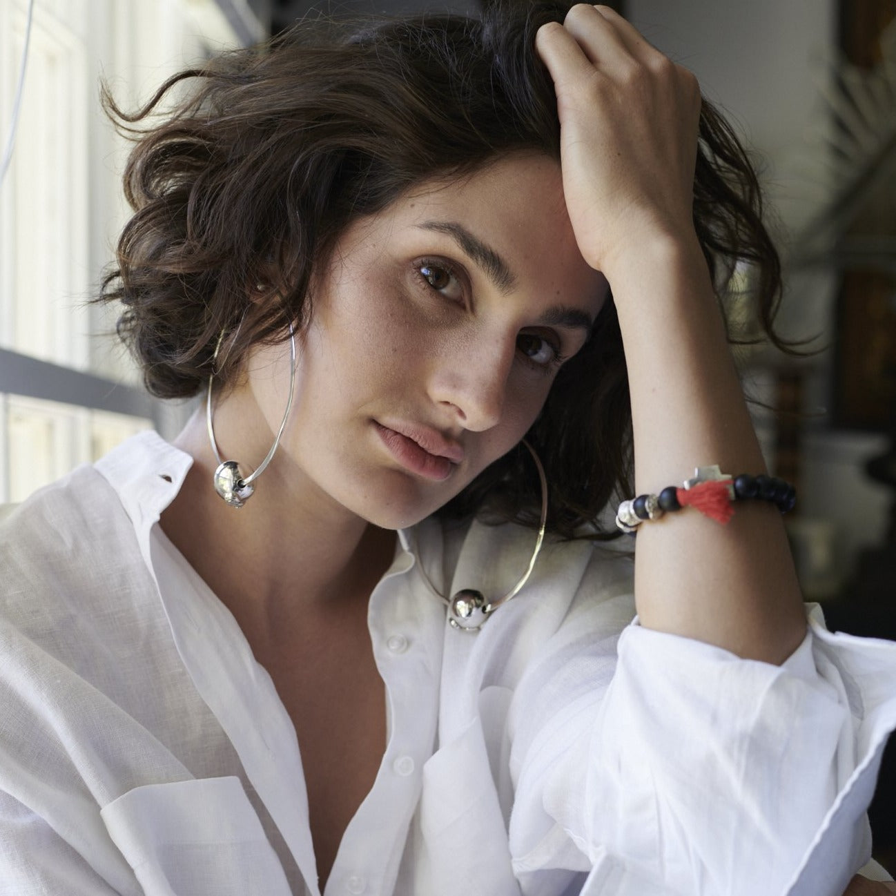 A short haired woman holding her head with her  hand. She is wearing a white shirt and two big silver hoop earrings with a silver bead in the center.