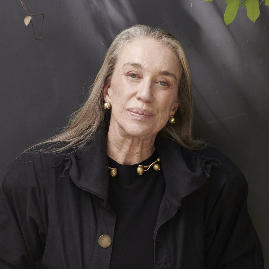 An older blonde woman resting agains a black background wearing black clothes and bronze jewelry.