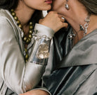 Two women close to each other. The hand of thee woman in the left is in the center. Both of them are wearing grey clothes and jewerly. The wrist in the center has a silver cuff with a clear cristal in the center.
