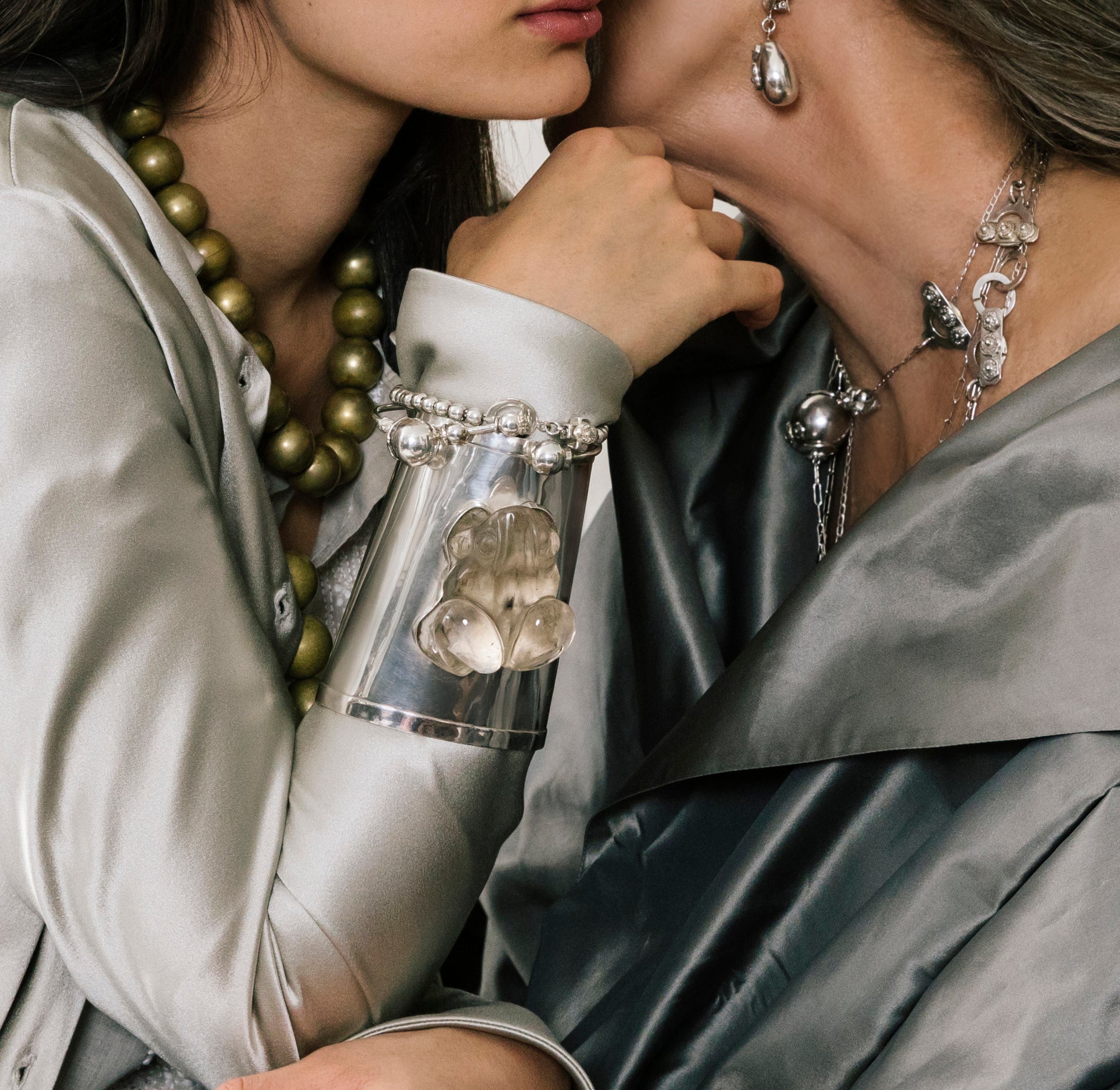 Two women close to each other. The hand of thee woman in the left is in the center. Both of them are wearing grey clothes and jewerly. The wrist in the center has a silver cuff with a clear cristal in the center.