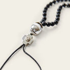A necklace with black beads and at the end a silver ball and a silver cross followed by two black leather strings. Plain white background.