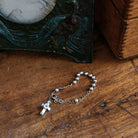 A delicate silver bead bracelet with a cross charm. The bracelet is resting in a wooden surface.