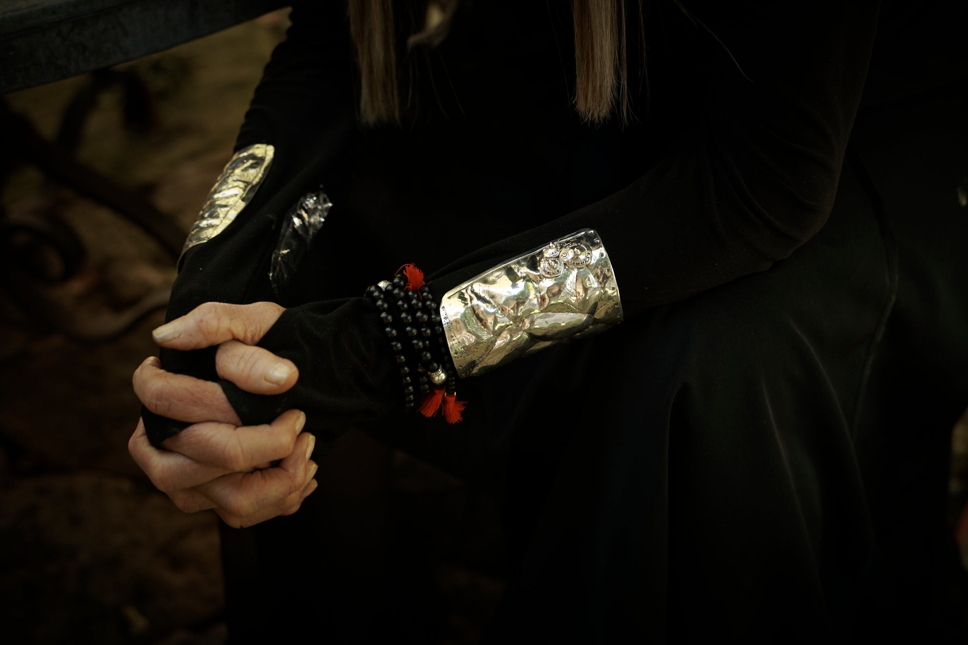 Hands of a woman clasp toguether. The woman is wearing black attire and silver cuffs with black beads bracelets.