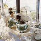 Three mouth blown puffed glass hearts with platinum finish and tiny iron rose on top of a white fabric. Surrounded by glasses.