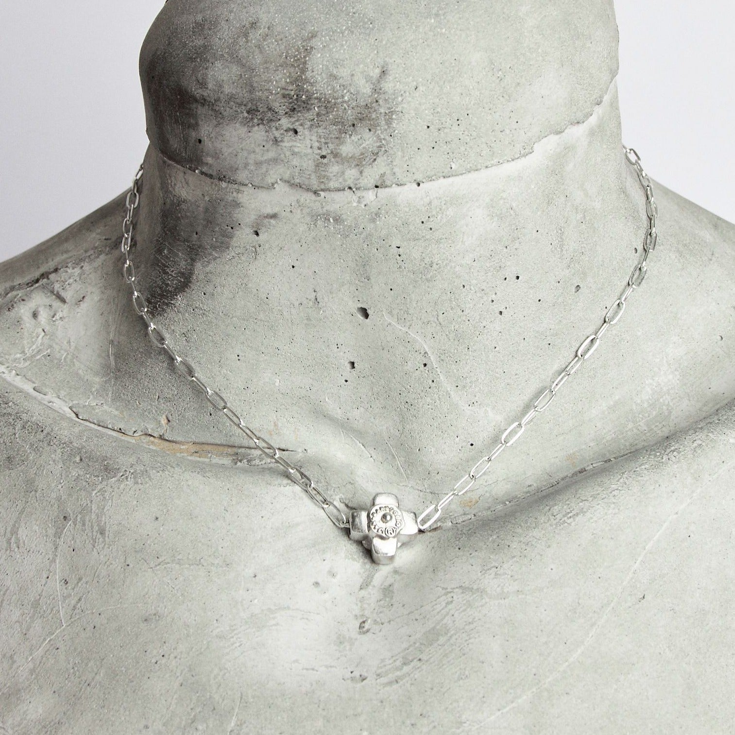 A delicate silver chain necklace with a silver cross with a silver seal with engraving.. The necklace is resting on the neck of a grey/ cement manikin.