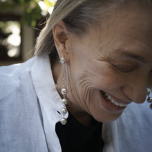 An older woman smiling and in the center is a silver beads hook earring. The background is blurry nature.