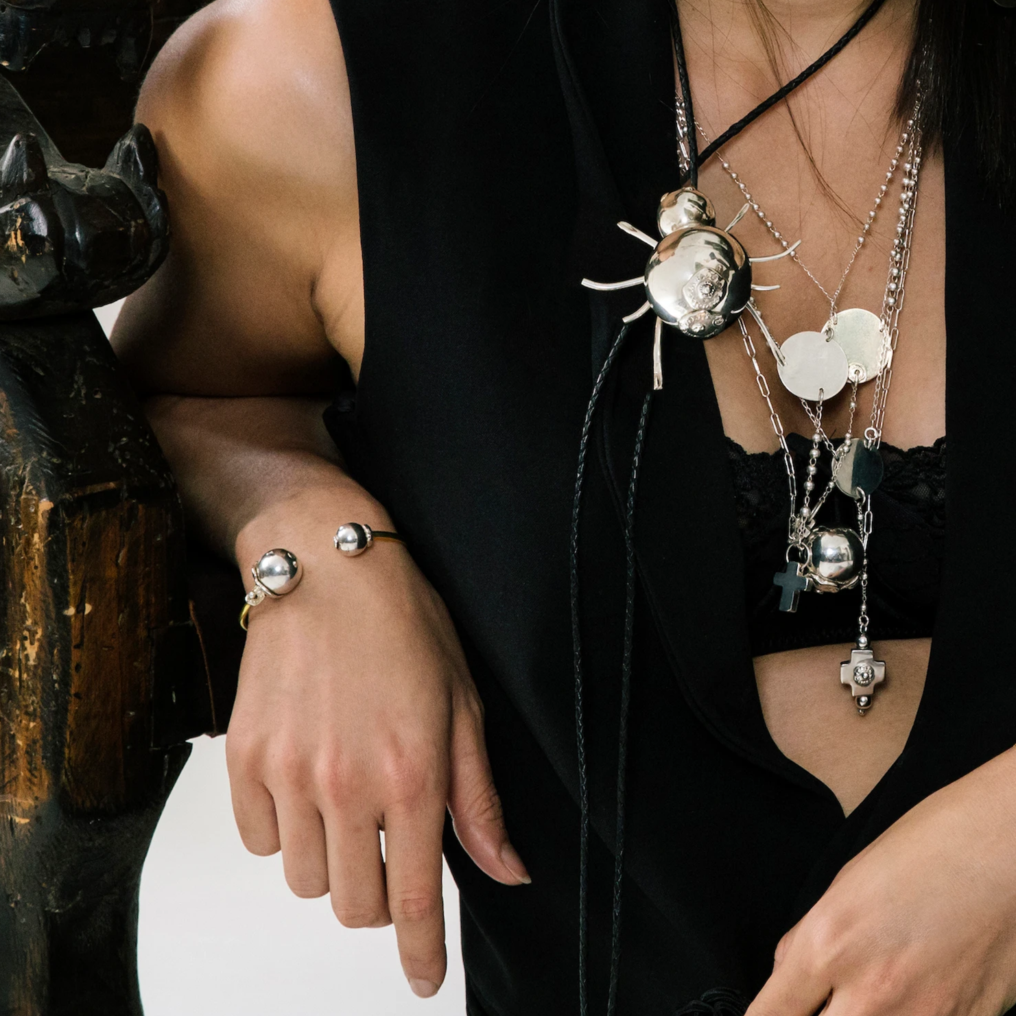 A torso of a woman  with black attire. Wearing several silver necklaces with crosses and spiders. In her wrist is a bracelet with silver studs.